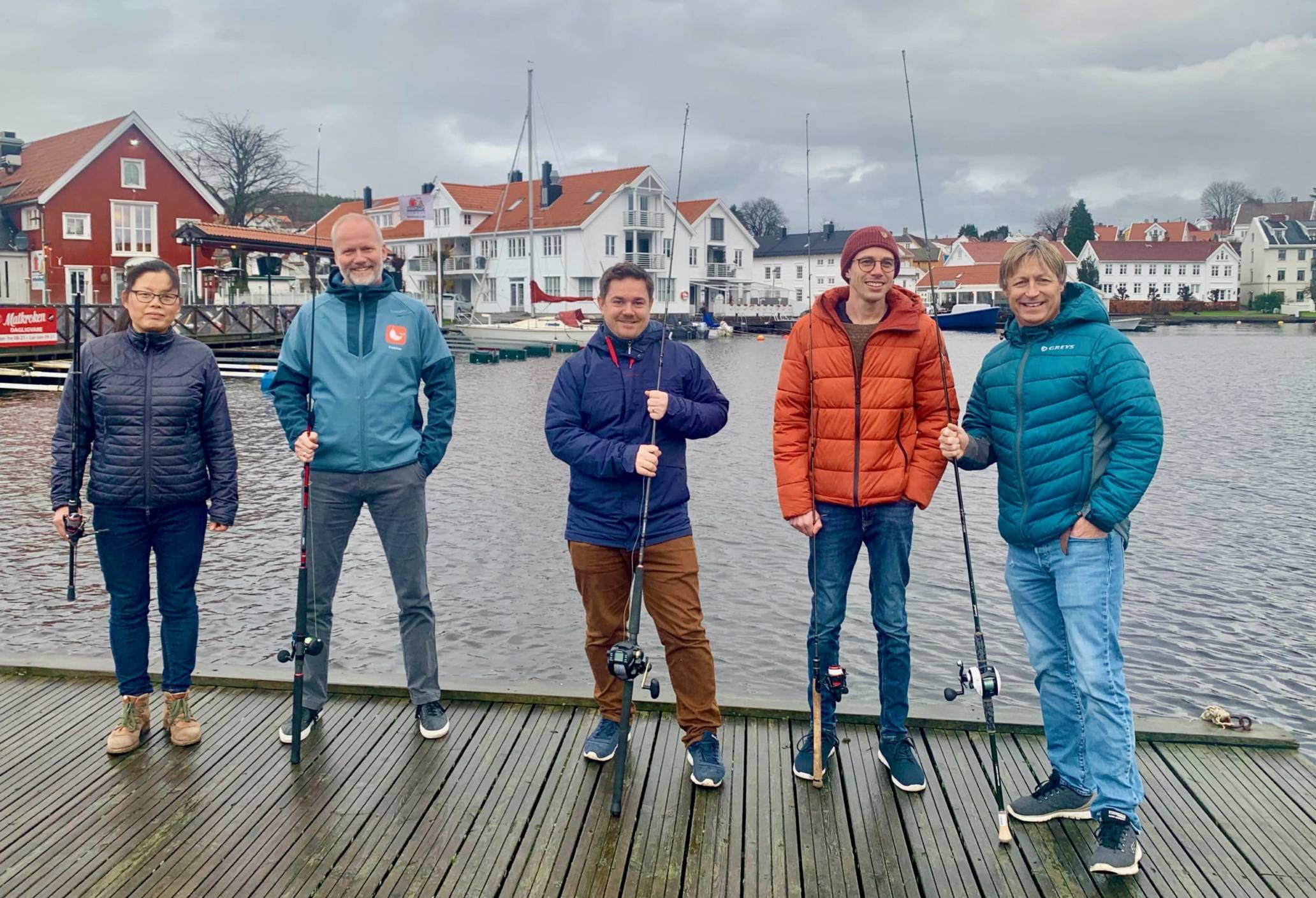 Fem glade personer med fiskestang i hånda på en flytebrygge foran et idyllisk havneområde