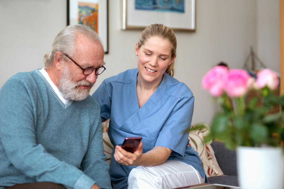 Helsepersonell sitter i sofa med eldre mann og viser han noe på mobileno